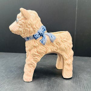 May include: A ceramic figurine of a lamb with a textured, curly cream-colored coat. The lamb features a painted blue ribbon tied in a bow around its neck. The figurine has a hollow opening on its back, indicating it functions as a small planter or vase.