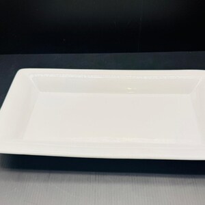May include: A rectangular white ceramic serving platter with a smooth, glossy finish and a wide, flat rim. The dish features a shallow basin and clean, straight edges, providing a simple and functional surface for presenting various food items.