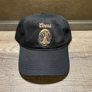 May include: A black baseball cap with a brown embroidered Coors logo featuring a mountain scene.