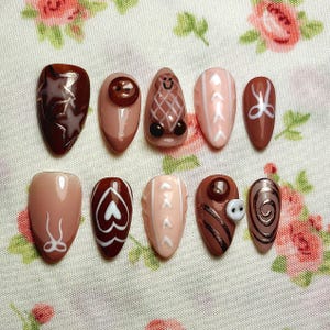May include: A collection of ten false nails in shades of brown and pink. Each nail is decorated with unique designs, including geometric patterns, hearts, and abstract art. Some nails have small decorative embellishments.