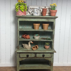 May include: A miniature green cabinet with shelves displays gardening-themed decor. Items include terracotta pots, a watering can, a goose figurine, and a sign that reads "POTTER'S HOME". The cabinet has two drawers and is set against a white wood-paneled wall.