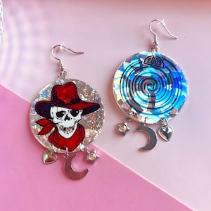 May include: A pair of dangle earrings. One earring features a skull wearing a red cowboy hat and bandana. The other earring has a blue and silver spiral design with the text "206S". Both earrings have silver hooks, charms, and a crescent moon.
