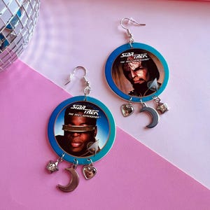 May include: A pair of Star Trek: The Next Generation earrings. Each earring has a blue and white circular design with a character image and the show's logo. Silver charms include a crescent moon, heart, and star.