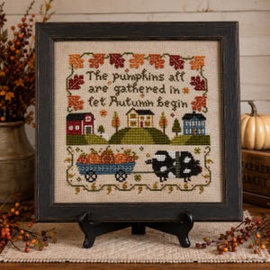 May include: A framed cross-stitch sampler on beige fabric featuring a rural landscape with a red barn, a house, a blue house, a wagon filled with pumpkins, and a black and white pig. The text reads: The pumpkins all are gathered in let Autumn begin.