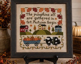 May include: A framed cross-stitch sampler on beige fabric featuring a rural landscape with a red barn, a house, a blue house, a wagon filled with pumpkins, and a black and white pig. The text reads: The pumpkins all are gathered in let Autumn begin.