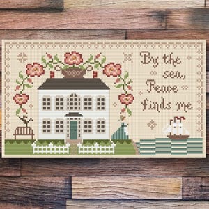May include: Cross-stitch pattern featuring a white house with a green door and a white picket fence. Pink flowers and vines surround the house. The text "By the sea, Peace finds me" is stitched in brown thread on a beige background.