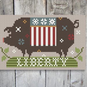 May include: Cross-stitch pattern featuring a brown pig wearing a red, white, and blue striped shirt with stars. The pig is standing on a green field with the word "LIBERTY" written below.