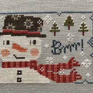 May include: Cross-stitch embroidery of a snowman wearing a brown and black hat with a red and white scarf. The snowman has a carrot nose and black eyes. The background is blue with white snowflakes and three green trees. The text "Brrr!" is stitched in blue.