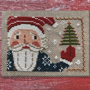 Op de afbeelding: Kruissteekontwerp met de Kerstman met een rode muts en want, die een groene kerstboom vasthoudt. Het kunstwerk is geplaatst tegen een neutrale achtergrond met sneeuwvlokken, perfect voor feestelijke decoraties.