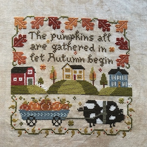 Könnte beinhalten: Kreuzstich-Stickerei mit einer Herbst-Szene mit einem Wagen voller Kürbisse, einer schwarz-weißen Kuh und drei Häusern. Der Text lautet "The pumpkins are gathered in, let Autumn begin."