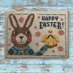 Op de afbeelding: Kruissteekontwerp met een bruin konijn dat een geel kuiken vasthoudt, met de woorden "HAPPY EASTER!" in marineblauw. Het konijn draagt een jasje met een teal en marineblauw patroon. Roze en gouden accenten maken het ontwerp compleet.