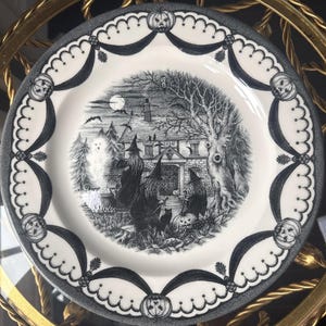 Könnte beinhalten: Ein runder Keramikteller mit einer schwarz-weißen Illustration von drei Hexen vor einem Haus, über dessen Tür die Jahreszahl 1866 eingraviert ist. Der Rand ist mit einem sich wiederholenden Muster aus Kürbissen und dunklen Girlanden verziert.