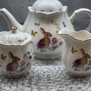 Puede incluir: Un juego de té de porcelana blanca con tetera, azucarero y jarrita para la leche. Cada pieza está decorada con un conejo marrón, mariposas y motivos florales. El juego tiene bordes dorados, añadiendo un toque de elegancia. Un juego de té con temática primaveral.