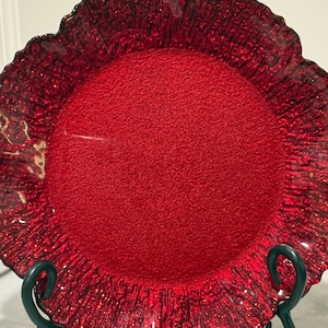 May include: A decorative red glass plate with a textured surface and a ruffled edge. The plate is round and sits on a dark green metal stand. The plate's color is a vibrant red.