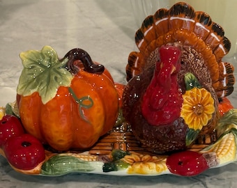Puede incluir: Un juego de salero y pimentero de cerámica que incluye una calabaza de color naranja brillante y un pavo marrón con carúncula roja, sobre una bandeja ovalada decorada con manzanas rojas, girasoles amarillos y mazorcas de maíz.