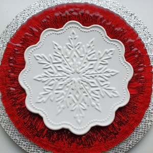 May include: A white snowflake-shaped plate sits atop a red, textured charger, which rests on a silver, glittery placemat. The plate has a scalloped edge and a raised snowflake design. The overall theme is festive and decorative.