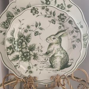 May include: Two white ceramic plates with a green floral and bunny design. The plates have scalloped edges and are displayed on a gold-colored metal stand. The plates feature a detailed illustration of a rabbit and floral patterns.