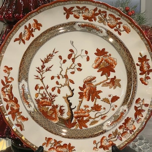 Puede incluir: Un plato de cena de cerámica blanca con un borde festoneado y un diseño floral en tonos naranja y marrón. El diseño presenta un árbol estilizado con ramas y hojas, y flores delicadas.