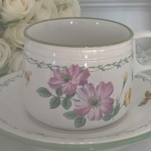May include: A white teacup and saucer with a floral design. The cup has a green rim and a floral pattern with pink and purple flowers, green leaves, and yellow accents. The saucer has a green rim and a floral pattern with green vines.