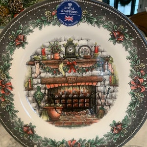Op de afbeelding: Een wit keramiek kerstbord met een zwarte en grijze rand. Het bord heeft een feestelijke scène van een open haard met kousen, een klok en een krans. Het bord is versierd met hulst, bessen en bellen. Het bord is gemaakt door Royal Stafford, een bedrijf dat in 1845 is opgericht. Het bord is gemaakt in Engeland.
