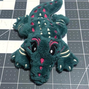 May include: A plush alligator toy in teal green with pink and white accents. The toy features a detailed design with embroidered eyes, a pink mouth, and pink spots along its back. The alligator is lying flat, showcasing its soft, textured fabric.
