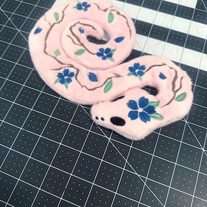 May include: A plush snake toy made of soft light pink fabric, coiled in a circular shape. The surface features embroidered brown branches, green leaves, and blue five-petaled flowers. The head has black embroidered eyes and nostrils.