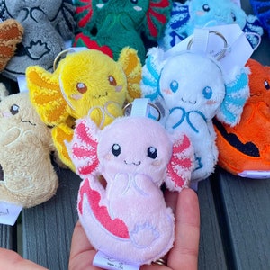 May include: A collection of plush axolotl keychains in various colors including pink, yellow, white, blue, green, and gray. Each soft, stuffed creature features embroidered facial details, prominent external gills, and a metal keyring attachment.