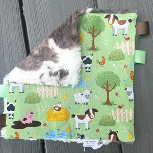 May include: A baby security blanket with a farm animal print on a green background. The blanket features cows, sheep, pigs, and chickens. The reverse side is a soft, gray and white plush material. It has brown and green ribbon tags.
