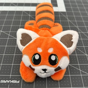 May include: A plush red panda toy with orange fur, a striped tail, and white and brown accents on its face and ears. The toy has large, round eyes and a smiling expression. The plush is lying on a gray cutting mat.