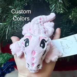 May include: A light pink plush alligator toy with embroidered black and white eyes, pink accents along the back, and a white tag attached. The text "Custom Colors" is visible in the upper left corner of the image, and the toy has a soft texture.