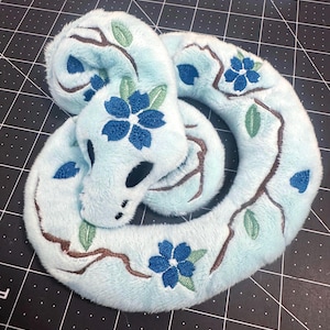 May include: A plush snake toy made of light blue fabric featuring embroidered brown branches, green leaves, and dark blue five-petaled flowers. The snake has solid black oval eyes and a coiled body shape with detailed stitching throughout the surface.