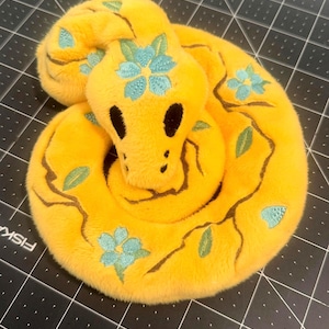 May include: A plush snake toy made of bright yellow fabric, coiled in a circle. The snake features embroidered brown vine patterns, light blue floral blossoms, and small green leaves. It has two large, solid black oval eyes and small black nostrils.