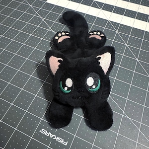 May include: A plush black cat toy with a soft, fuzzy texture. The cat features large, embroidered eyes with green irises, light pink inner ears, and small pink paw pads on its feet. The design is stylized with a rounded head and a long tail.