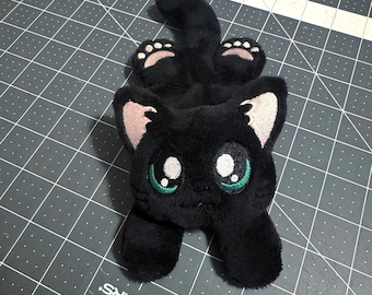 May include: A plush black cat toy with a soft, fuzzy texture. The cat features large, embroidered eyes with green irises, light pink inner ears, and small pink paw pads on its feet. The design is stylised with a rounded head and a long tail.