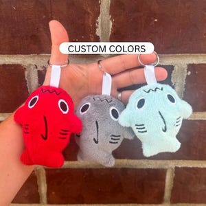 May include: Three plush shark keychains in red, gray, and light blue. Each shark has white eyes, a black mouth, and a white ribbon loop. The text "CUSTOM COLORS" is above the keychains.