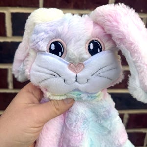 May include: A pastel tie-dye plush bunny with embroidered eyes, nose, and whiskers. The bunny has long, floppy ears and a soft, cuddly texture.