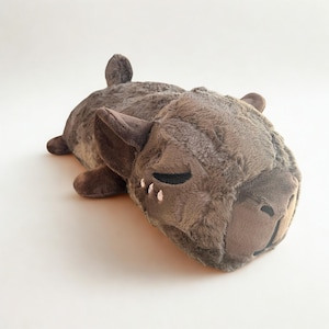 May include: A plush, brown stuffed animal in the shape of a sleeping creature. The toy has dark brown ears, a snout, and closed eyes with black and white details. The soft toy is lying down on a white surface.