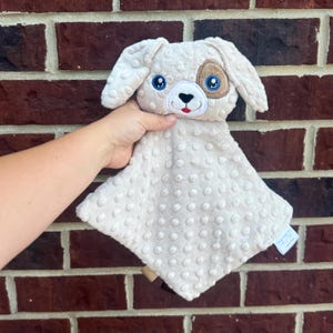 May include: A beige dog-shaped baby security blanket with a textured, dotted surface. The dog has blue eyes, a brown eye patch, and a red mouth. The blanket is held against a brick wall.