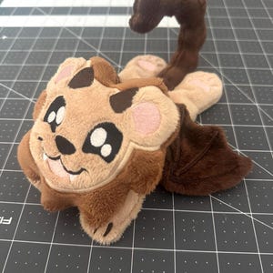 May include: A plush toy featuring a tan face with dark brown accents, large black and white embroidered eyes, and a small smile. The creature has dark brown bat-like wings, a long curled tail, and light tan paws with pink embroidered pads.