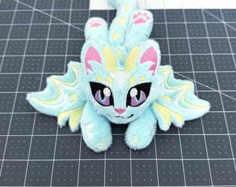 May include: A plush toy in the shape of a fantasy creature, primarily light blue with yellow and pink accents. The creature has large eyes, wings, and a tail. The toy is lying on a gray cutting mat.