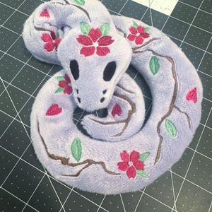 May include: A plush snake toy in a coiled position, featuring a soft light purple fabric. The surface is decorated with embroidered brown branches, green leaves, and vibrant pink cherry blossom flowers. The snake has solid black oval eyes.