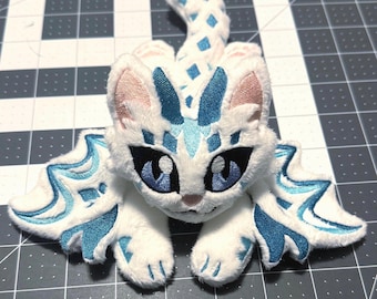 Op de afbeelding: Een witte pluche kat met blauwe en lichtblauwe geborduurde details. De kat heeft grote blauwe ogen, roze binnenoren en een staart met blauwe ruitpatronen. De vleugels en de kop hebben ingewikkelde blauwe ontwerpen.