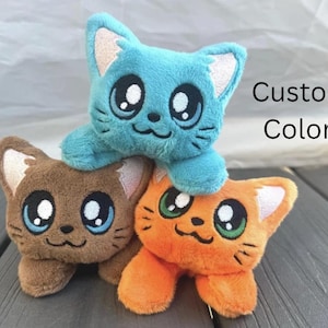 May include: Three plush cat toys in blue, brown, and orange, stacked together. Each cat has large, expressive eyes and embroidered details. The text "Custom Colors" is visible in the background.