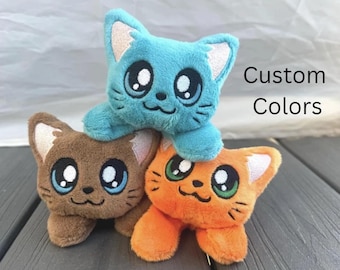 May include: Three plush cat toys in blue, brown, and orange, stacked together. Each cat has large, expressive eyes and embroidered details. The text "Custom Colors" is visible in the background.