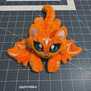 May include: A plush orange cat-like creature with large teal eyes, pointed ears, and two sets of wings extending from its sides. The creature features embroidered white markings on its forehead, tail, and wings, with a soft, fuzzy texture.