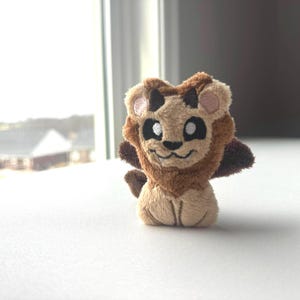 May include: A small, plush lion toy with a friendly expression. The lion has a tan body, a brown mane, and small, dark horns. Its face features large, white eyes, a black nose, and a smiling mouth. The toy is sitting on a white surface.