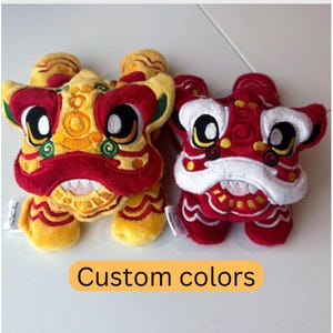 May include: Two plush lion dance toys. One is yellow and red, the other is red and white. Both have large eyes and embroidered details. The text "Custom colors" is in an orange rectangle below the toys.