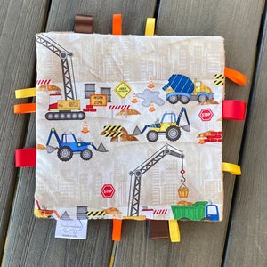 Op de afbeelding: Een beige babydeken met een bouwthema. De deken heeft verschillende bouwvoertuigen, waaronder een kraan, een betonmixer, een bulldozer en een graafmachine. De deken heeft ook verschillende kleurrijke stoffen labels aan de randen.
