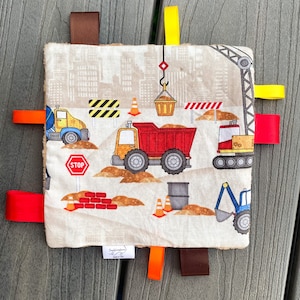 Op de afbeelding: Een beige babydeken met een bouwthema. De deken heeft een rode vrachtwagen, een blauwe vrachtwagen, een gele kraan en een stopteken. De deken heeft verschillende kleurrijke stoffen banden.