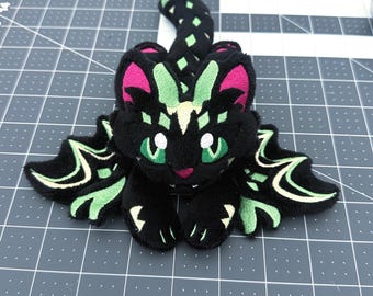 Op de afbeelding: Een zwarte pluche kat met groene ogen, roze oren en groene en crèmekleurige accenten op de vleugels en staart. De kat ligt op een grijs oppervlak met een rasterpatroon.